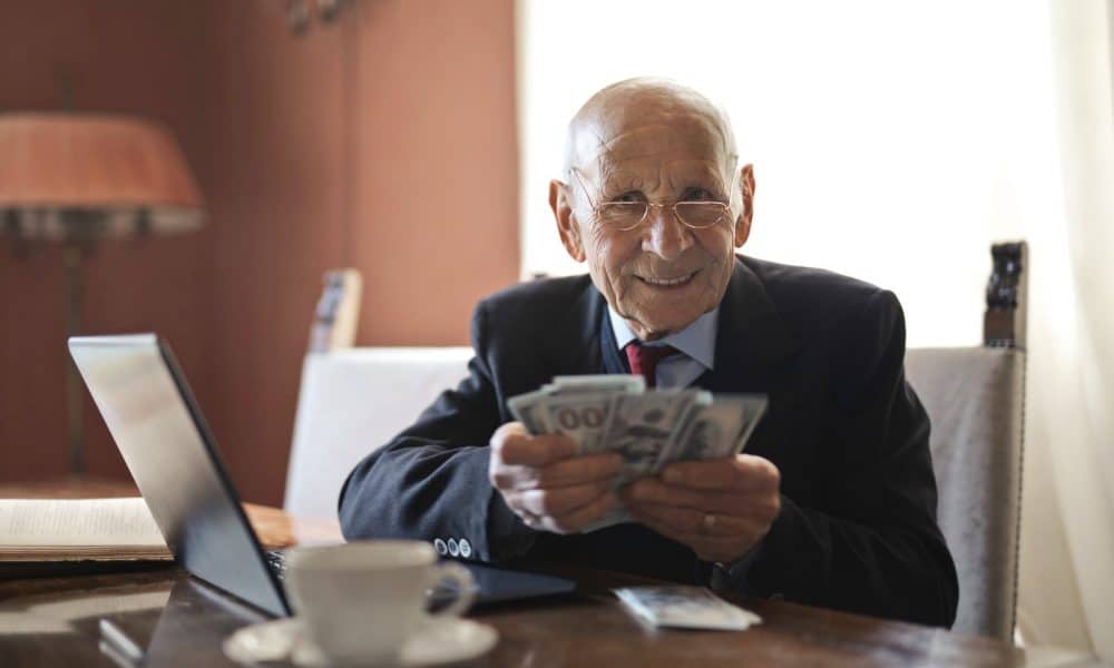 Confident senior businessman holding money in hands while sitting at table near laptop
