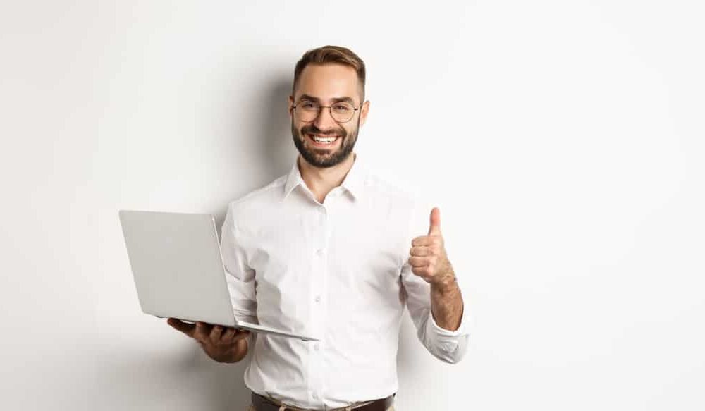 business young successful male entrepreneur showing thumbup while working on laptop standing over wh