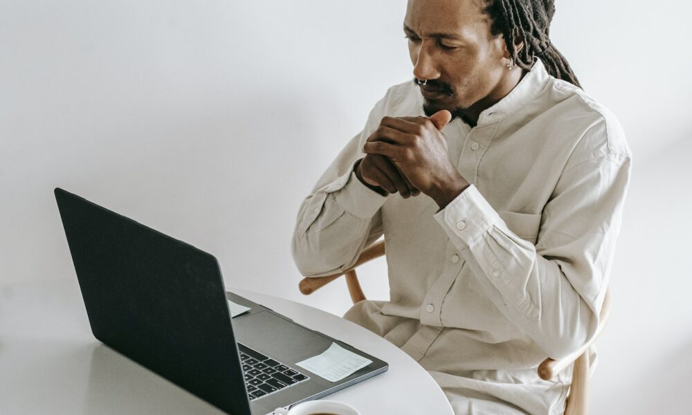 Concentrated adult African American male in formal wear sitting at table with cup of coffee and watching video on netbook in light room