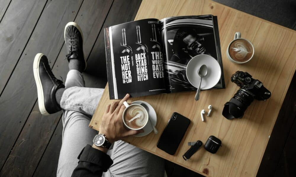 Top View Of A Table With Man Holding A Cup Of Latte With A Magazine, Camera, Cellphone And Other Personal Effects