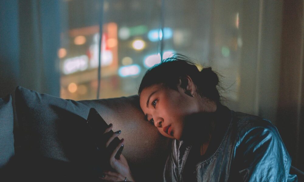 A woman looking at her mobile phone at night with city lights blurred in the background.