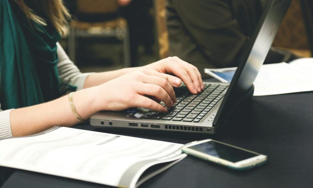 Adult woman typing on a laptop at a desk with a smartphone and documents.