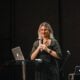 A woman giving a presentation on stage with a microphone and laptop.
