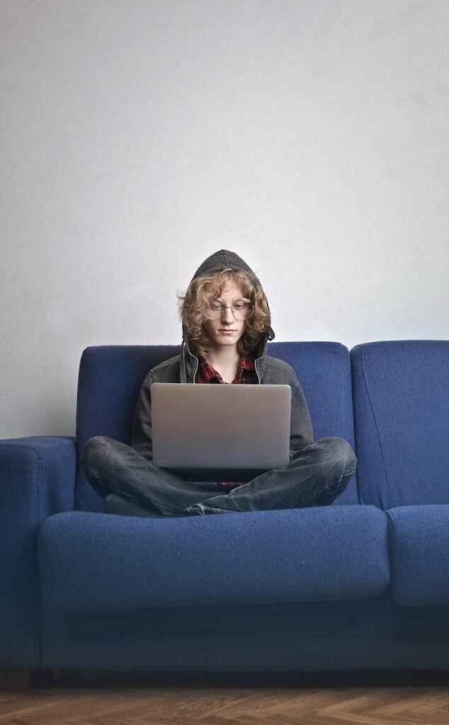 Person with glasses typing on laptop while sitting cross-legged on a blue couch.