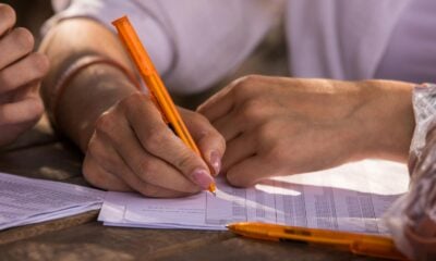 Close-up shot of hands writing on documents with orange pen, implying business or education setting.