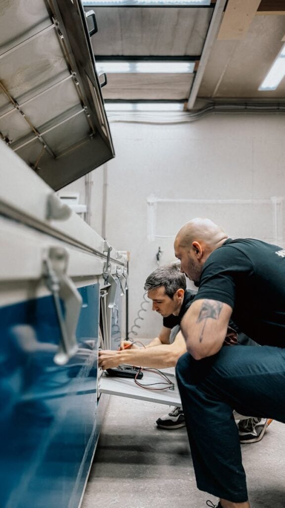 Two technicians repairing a machine indoors, displaying teamwork and focus.