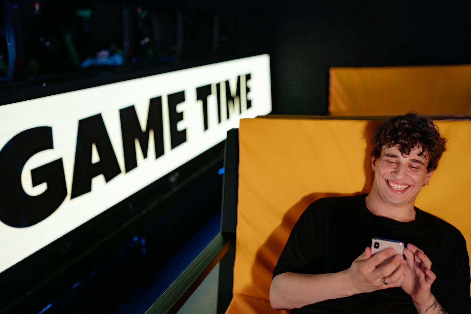 Smiling young man gaming on smartphone in a cozy indoor setting with 'Game Time' sign.