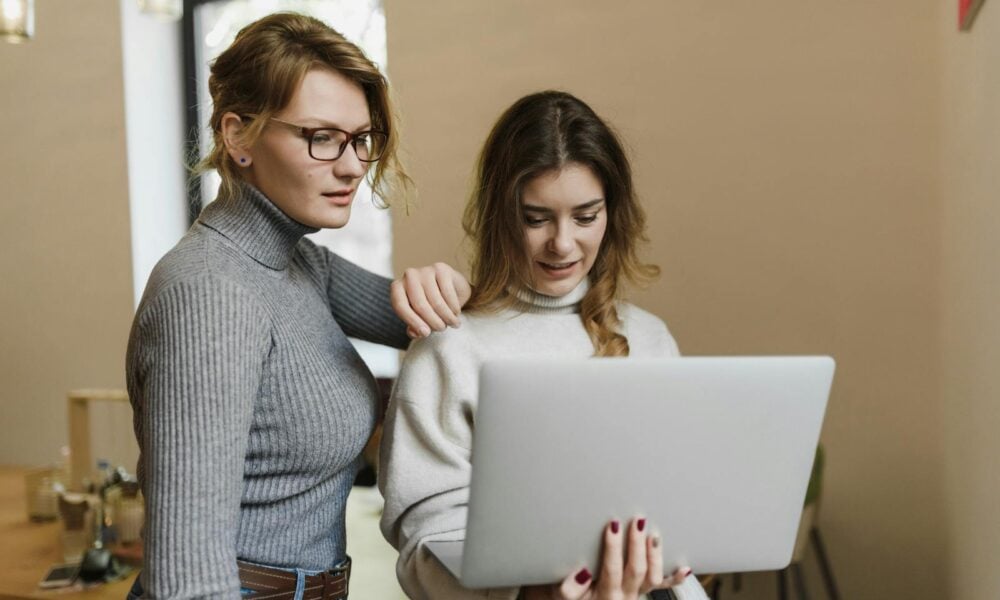 Two women in turtlenecks work together on a laptop in a warm indoor setting, showcasing teamwork.