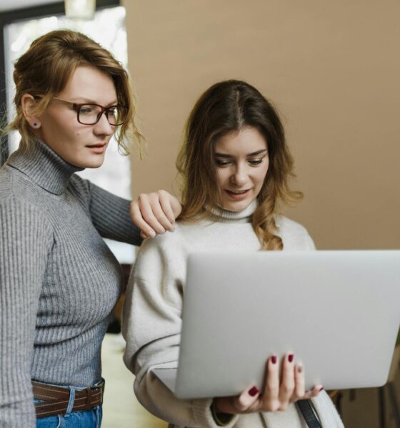 Two women in turtlenecks work together on a laptop in a warm indoor setting, showcasing teamwork.