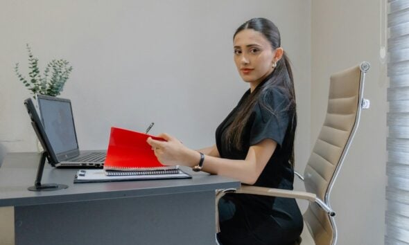 Woman working at desk with laptop and red folder.