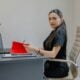 Woman working at desk with laptop and red folder.