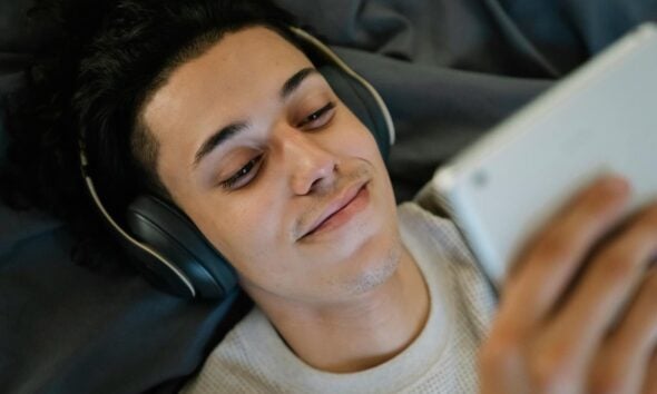 Positive young Hispanic man in headphones smiling and watching film on tablet while relaxing on bed at home
