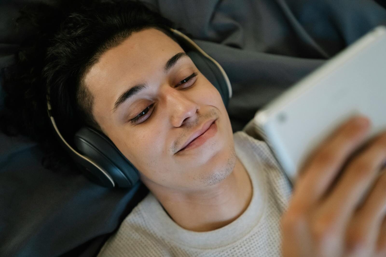 Positive young Hispanic man in headphones smiling and watching film on tablet while relaxing on bed at home