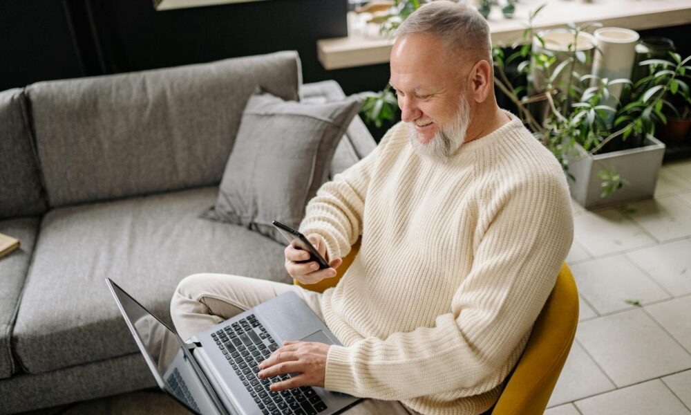 A smiling elderly man sits comfortably indoors, using both a phone and laptop, with a cozy ambiance.
