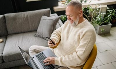 A smiling elderly man sits comfortably indoors, using both a phone and laptop, with a cozy ambiance.