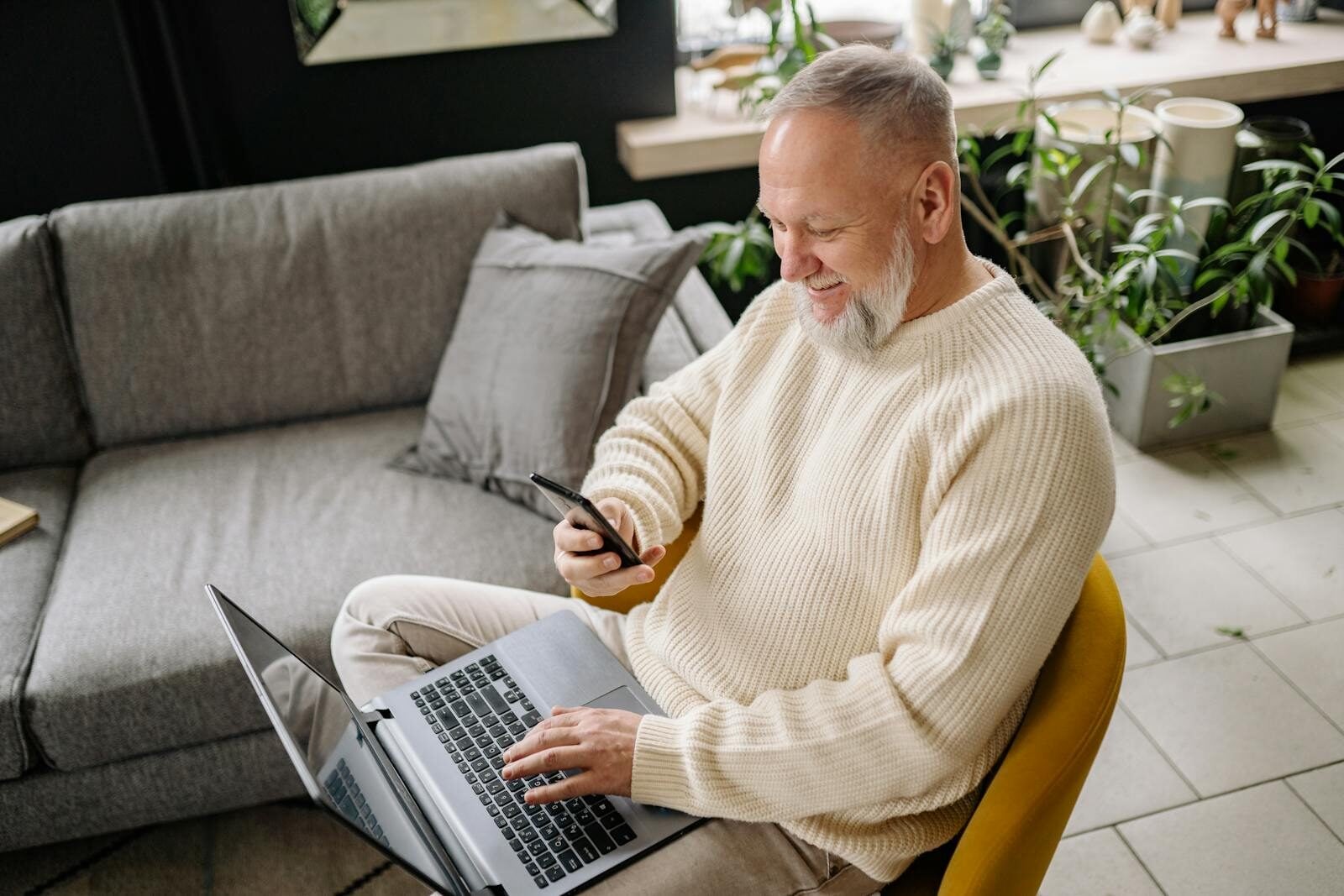 A smiling elderly man sits comfortably indoors, using both a phone and laptop, with a cozy ambiance.