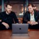 two men sitting at a table looking at a laptop