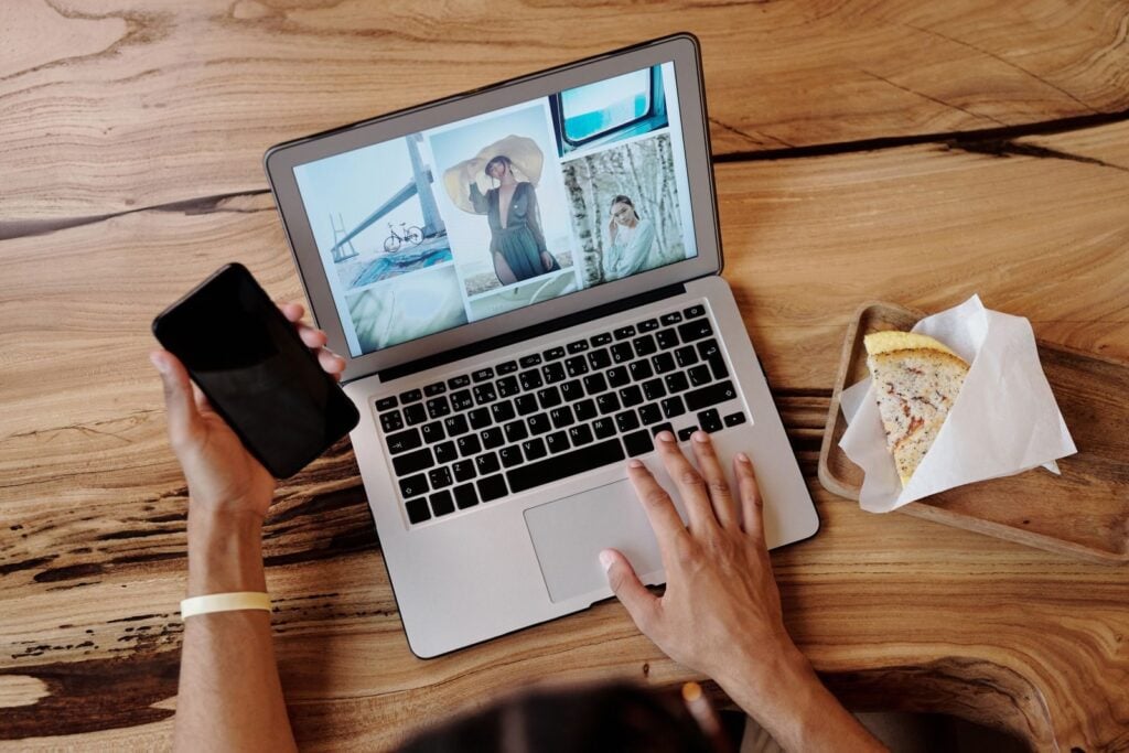 Person uses smartphone and laptop while enjoying a sandwich at a wooden table, showcasing multitasking and technology use.
