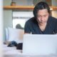 An Asian man working from home on a laptop, focusing on his digital tasks in a cozy indoor setting.