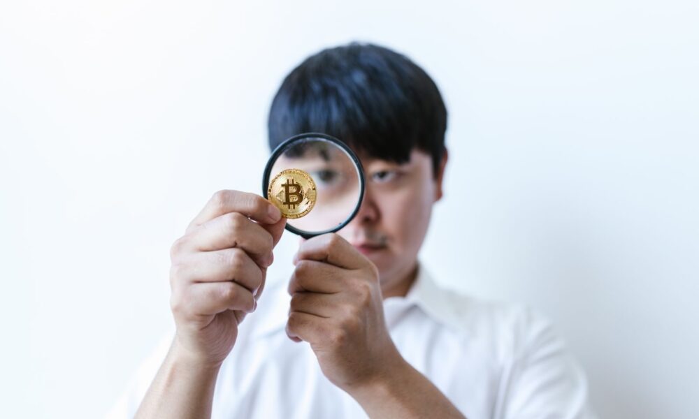 Close-up of a man examining a Bitcoin coin with a magnifying glass, symbolizing cryptocurrency analysis.