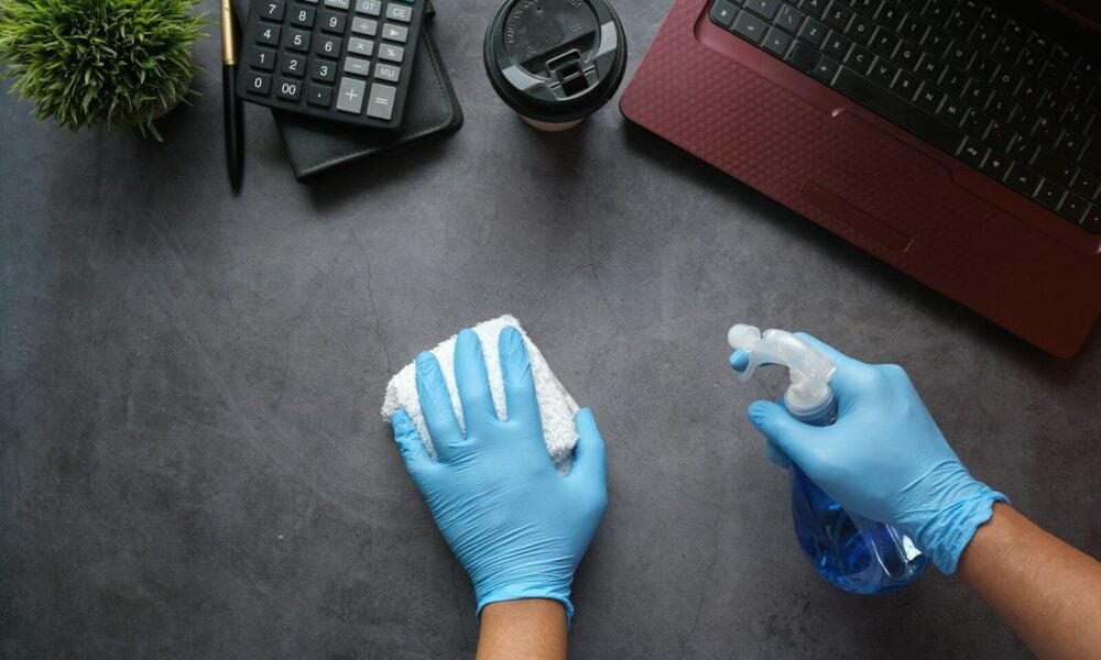 Hands in blue gloves cleaning a dark desk with a spray bottle and cloth