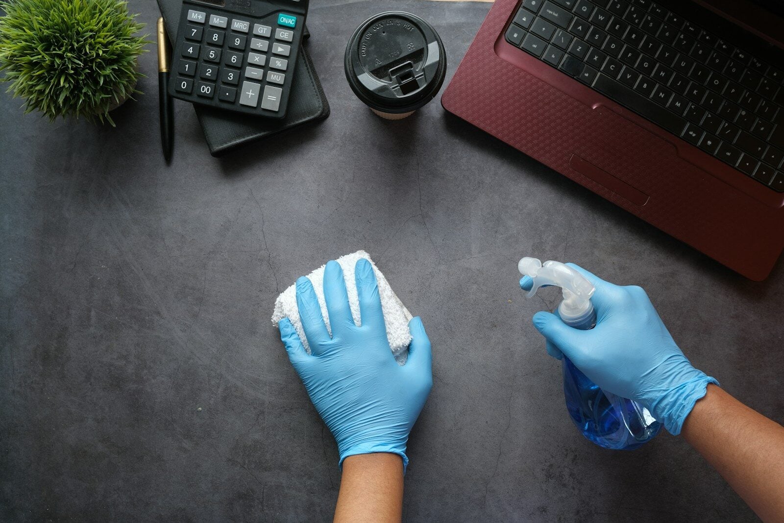 Hands in blue gloves cleaning a dark desk with a spray bottle and cloth