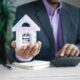 man holding model house at desk with calculator