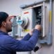 A person in a blue hoodie and baseball cap working on an electrical meter