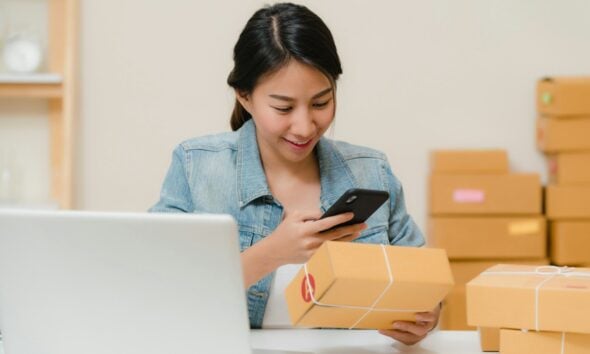 Woman checking package with phone near laptop and boxes