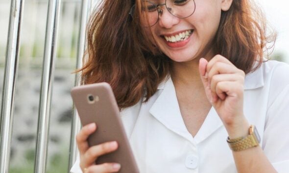 A woman in a white shirt laughing while looking at her smartphone