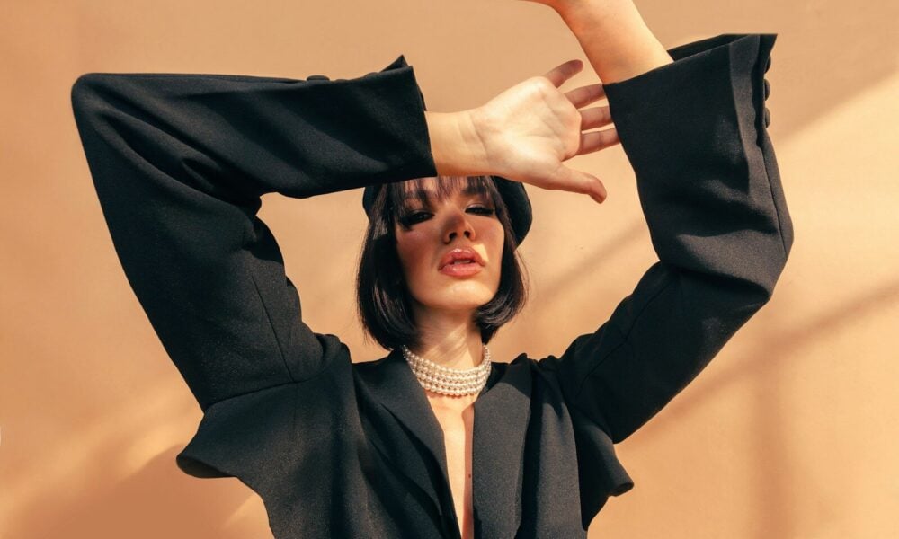 A woman in a black blazer and pearl necklace posing against a tan wall