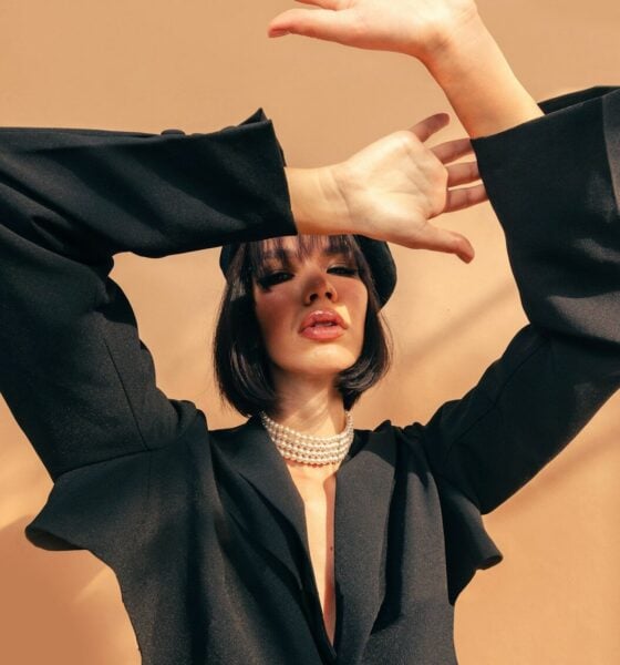 A woman in a black blazer and pearl necklace posing against a tan wall