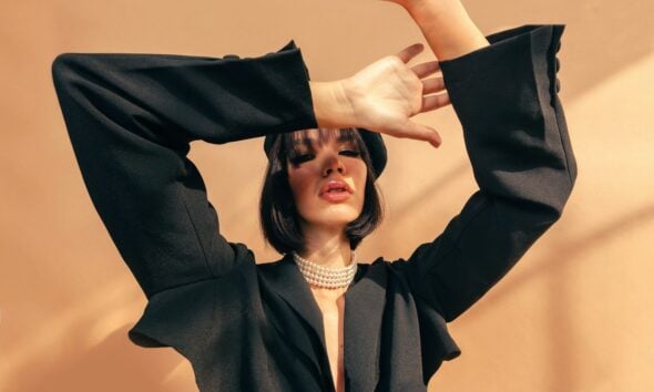 A woman in a black blazer and pearl necklace posing against a tan wall