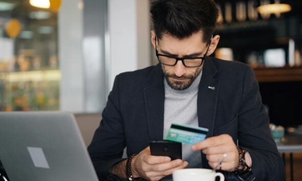 a man sitting at a table looking at his cell phone and holding a credit card