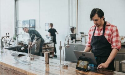 A barista in a plaid shirt and apron using a tablet at a wooden counter