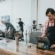 A barista in a plaid shirt and apron using a tablet at a wooden counter